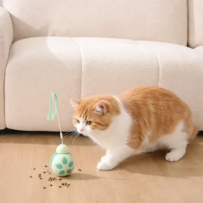 An orange and white ginger cat pouncing toward a green treat dispenser toy with kibble scattered on the floor.