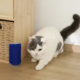A grey and white cat with bright eyes crouches on a wooden floor, pawing and rubbing its chin against a detached blue Cat Self-Groomer brush. The cat's long, dark tail is curved gracefully behind it. A woven basket and cabinet are in the background.