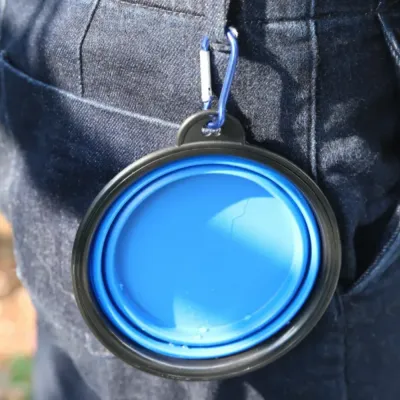 A blue collapsed silicone dog bowl clipped to a person's denim jeans using a blue metal carabiner.