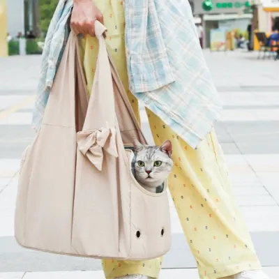 A person carrying a grey tabby cat in a beige shoulder pet tote bag while walking outdoors.