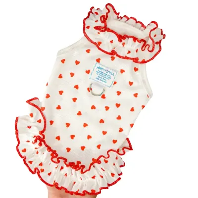 red-heart-pattern-dog-harness-dress-with-ruffles Close-up of a white dog dress with red heart prints, featuring a ruffled collar and a built-in metal D-ring for leash attachment.