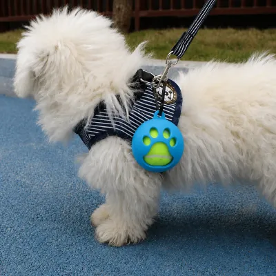 Portable Dog Tennis Ball Holder with Paw Shape for Outdoor Walking Fluffy white dog wearing a striped harness with a blue paw-shaped silicone tennis ball holder attached during an outdoor walk