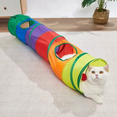 White cat resting inside a colorful rainbow cat tunnel with side openings and a hanging red pom-pom toy.