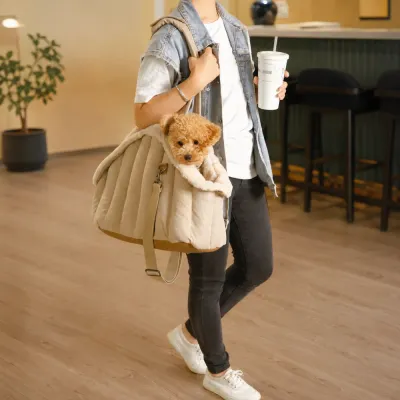 Person carrying a small dog in a beige plush dog carrier bag with shoulder handles inside a modern café.