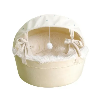 Cream enclosed cradle cat bed with plush lining, canopy, bow ties and hanging pom-pom toy on white background